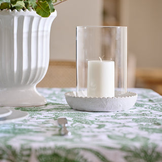 Clear candleholder with decorative green base on a table with a decorative cloth and plant