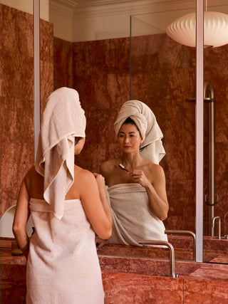 Woman applying makeup in a bathroom with towels on her head