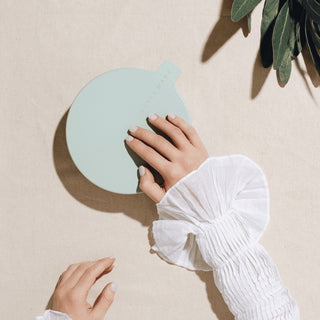Hand holding a light blue round mirror on a beige surface with a plant shadow.