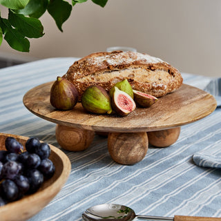 Loaf of bread with figs on a wooden stand on a table with a bowl of grapes and a spoon.