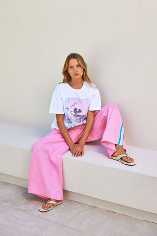 Woman sitting on a white ledge wearing a white t-shirt with a colorful graphic and pink pants.