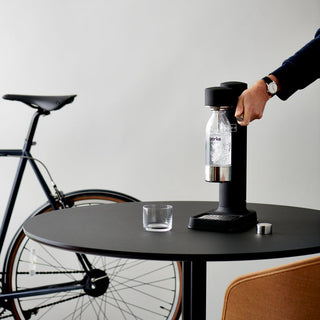 Person using a black carbonation device on a table with a bicycle in the background.
