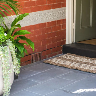 Front door entrance with a doormat, plant, and brick wall.